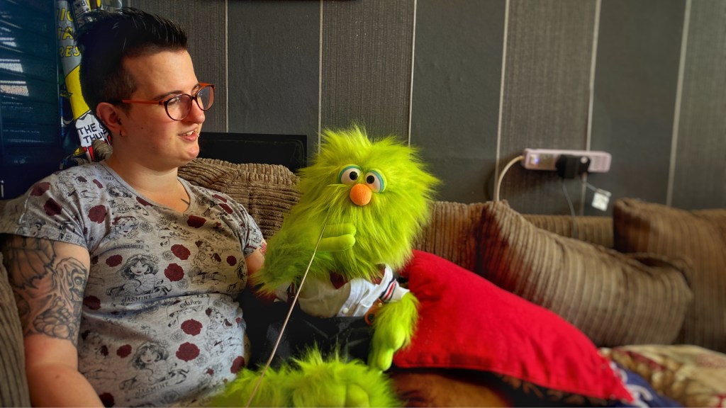 A woman practicing ventriloquy with a green furry puppet.  