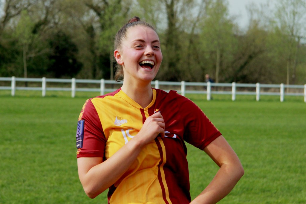 Ash has brown hair tied up in a high pony tail. She is clutching the Bradford city logo on her top whilst laughing