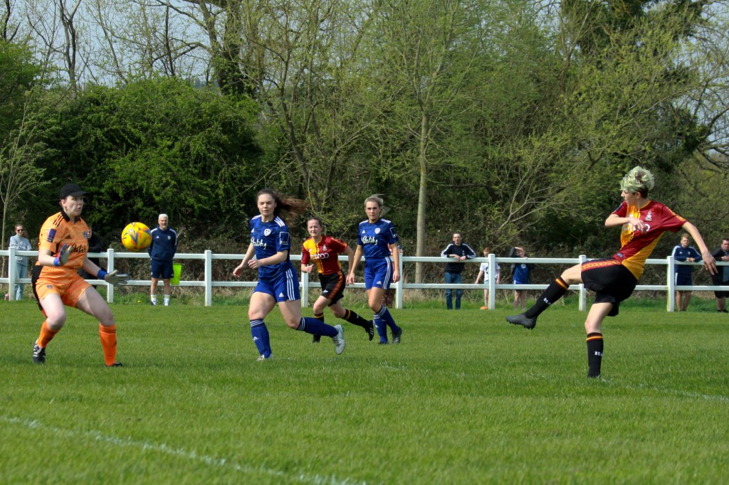 Val on the right side of the frame take a shot. The ball is travelling towards the goalkeeper who tries to save. Three players are running in the background whilst spectators watch