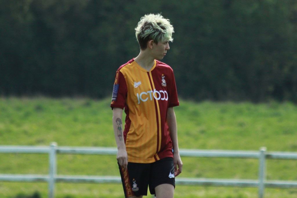 Val stands alone on the pitch with her head turning right. She is wearing her claret and amber football kit. She has short hair which is washed out blue colour. she also has a tattoo on her arm of an anime character