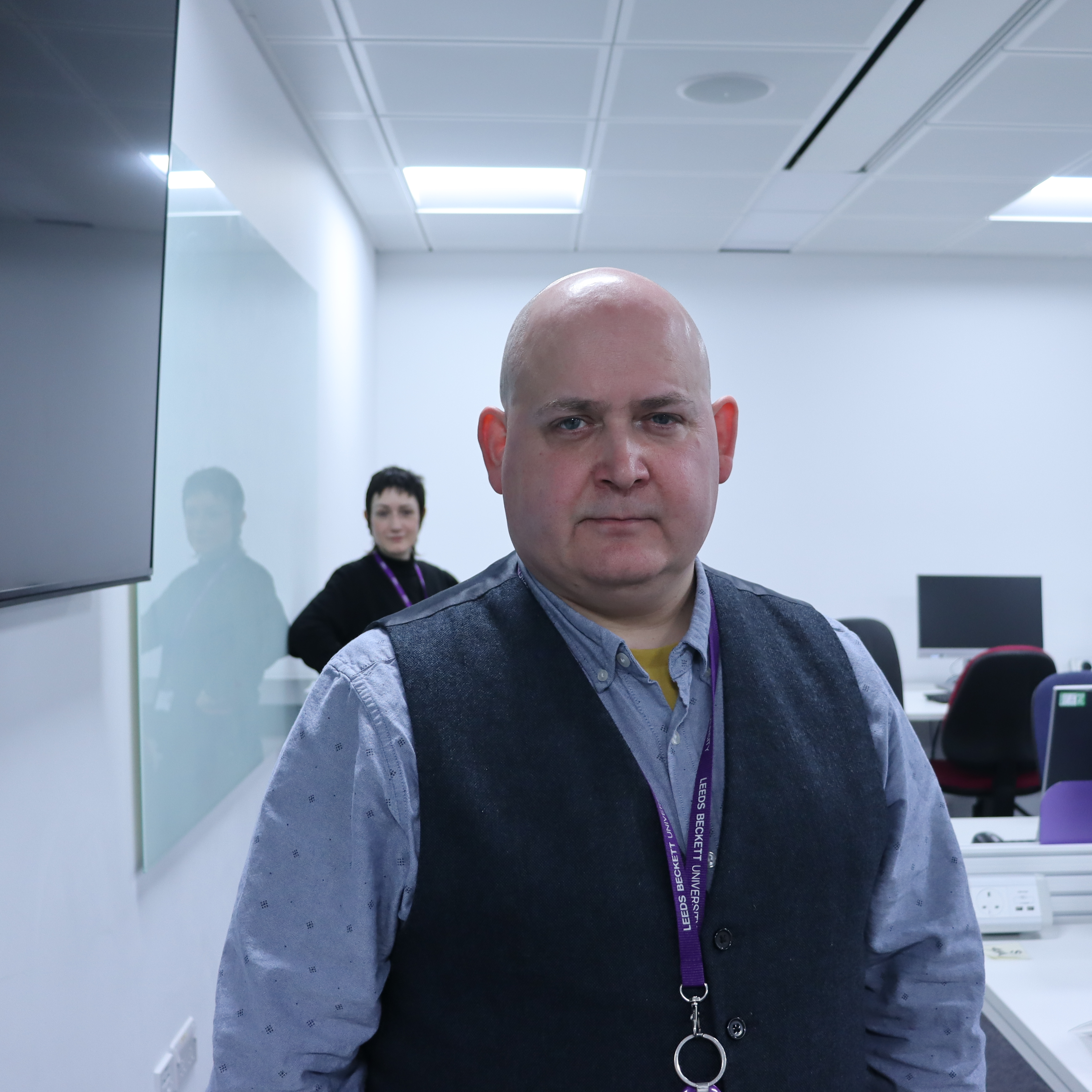 An image of a man looking at the camera, with a female stood behind him. The female isn't in focus.