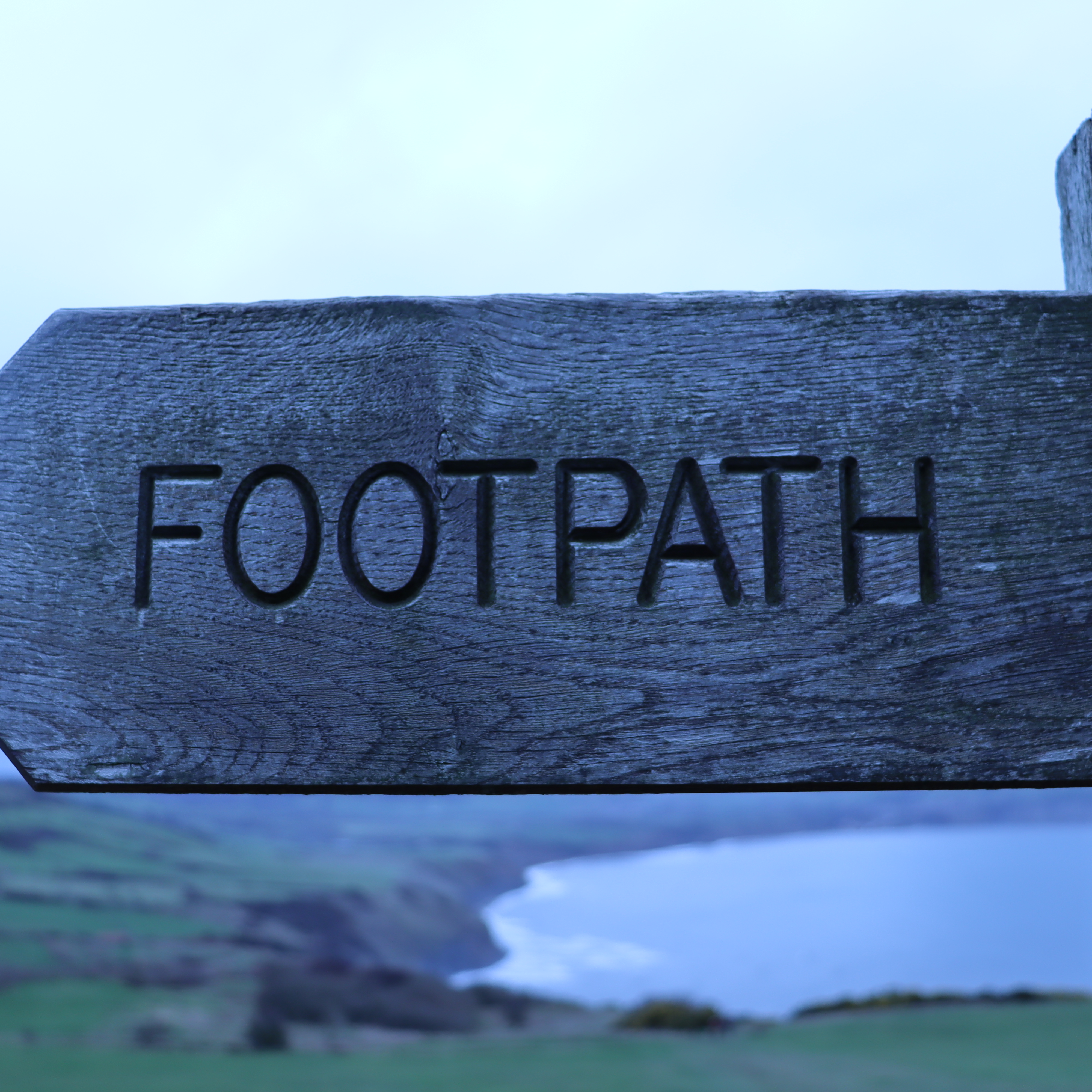 A wooden sign saying footpath with a blurry background of cliffs meeting the ocean.