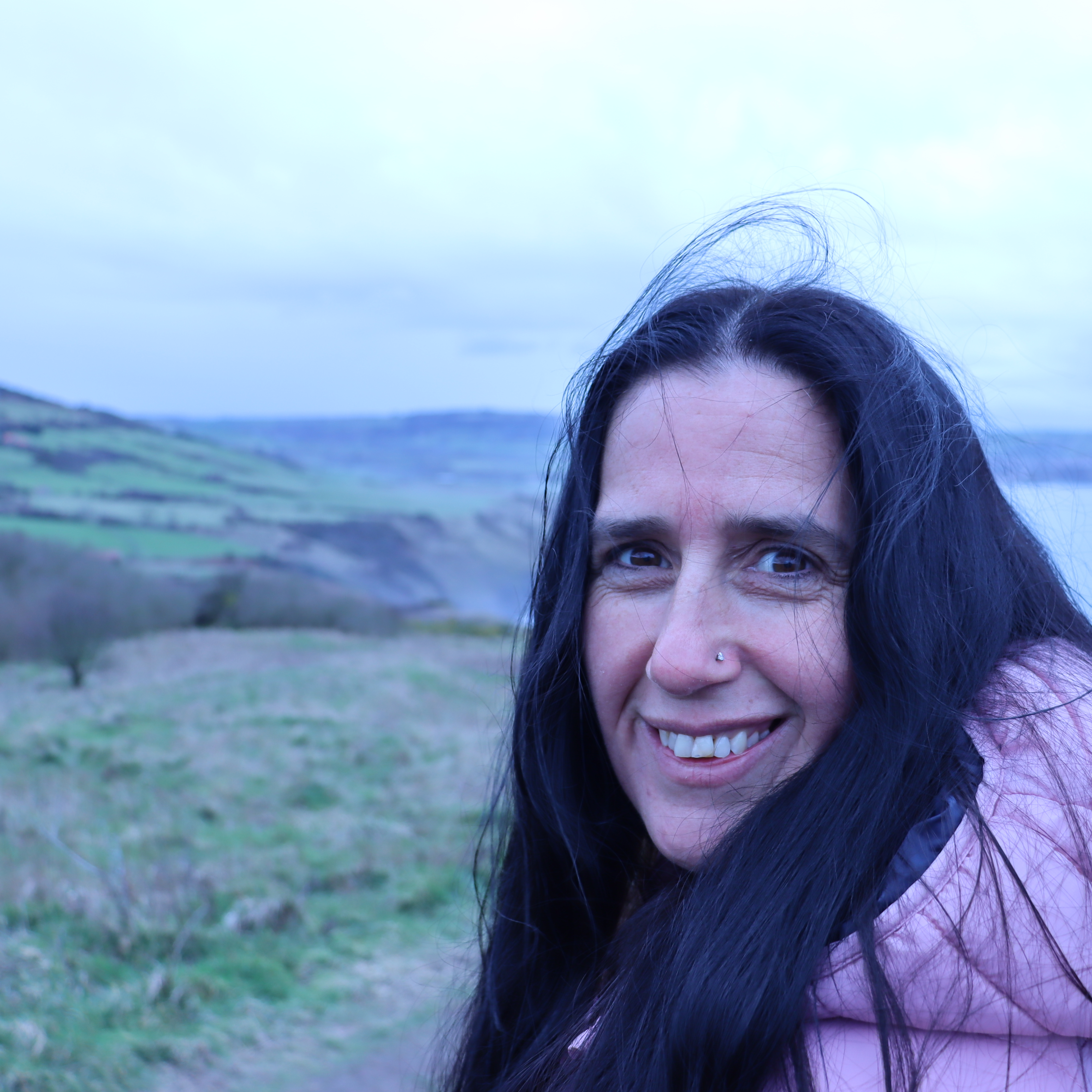 An image of a woman smiling with black hair wearing a pink coat, with the wind blowing her hair. With cliffs and the ocean behind her that are blurry.