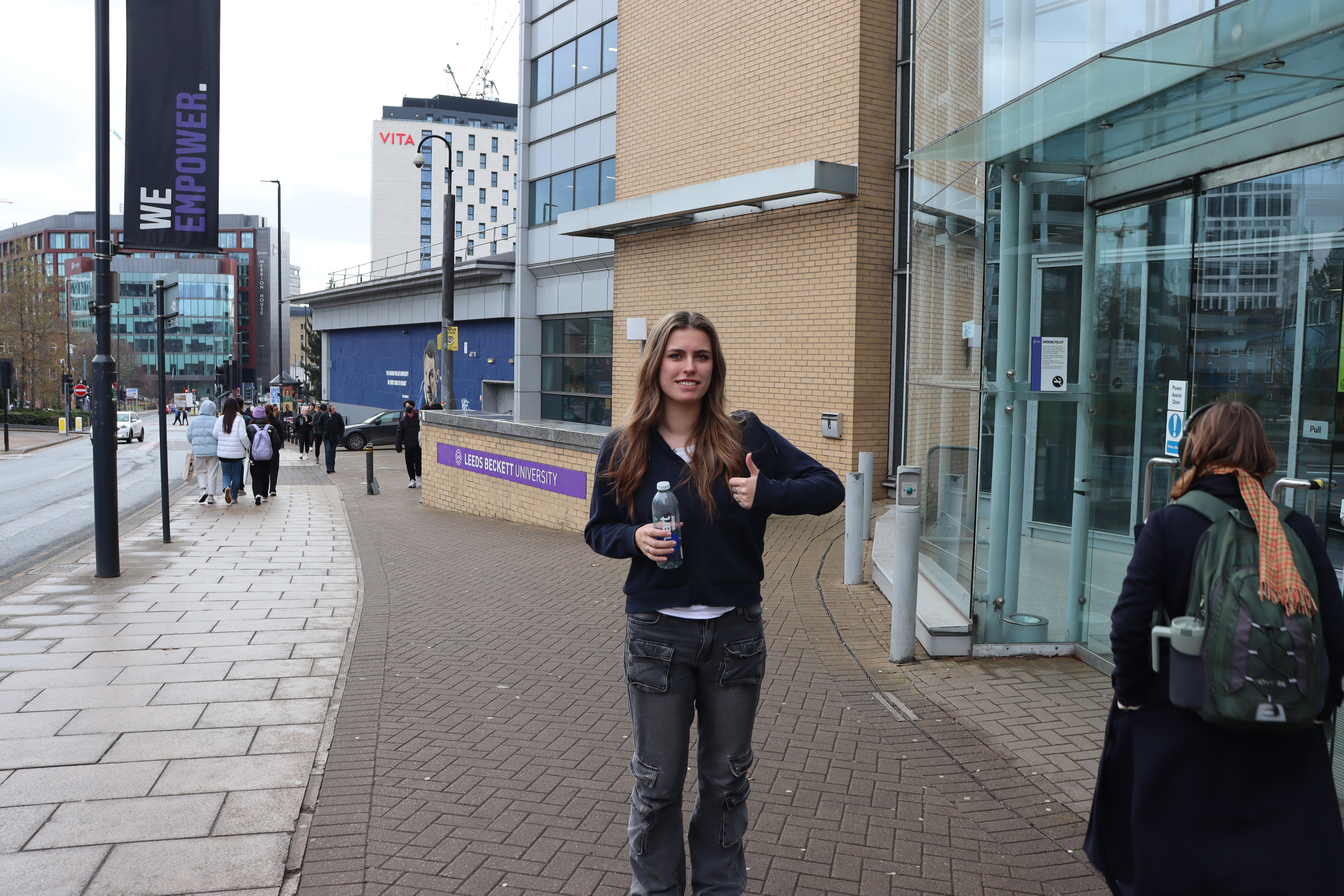 An image of a woman looking at the camera, holding a water bottle with her thumb up.