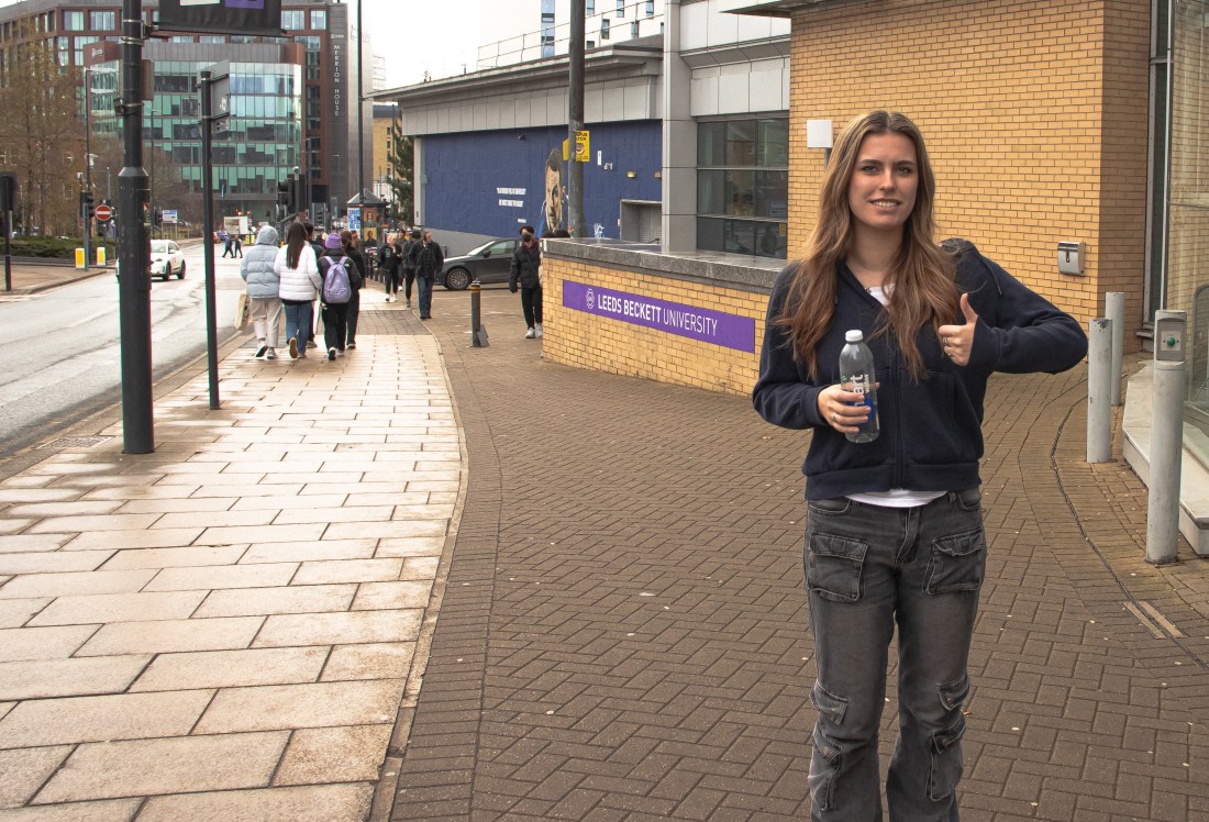 An image of a woman looking at the camera, holding a water bottle with her thumb up.
