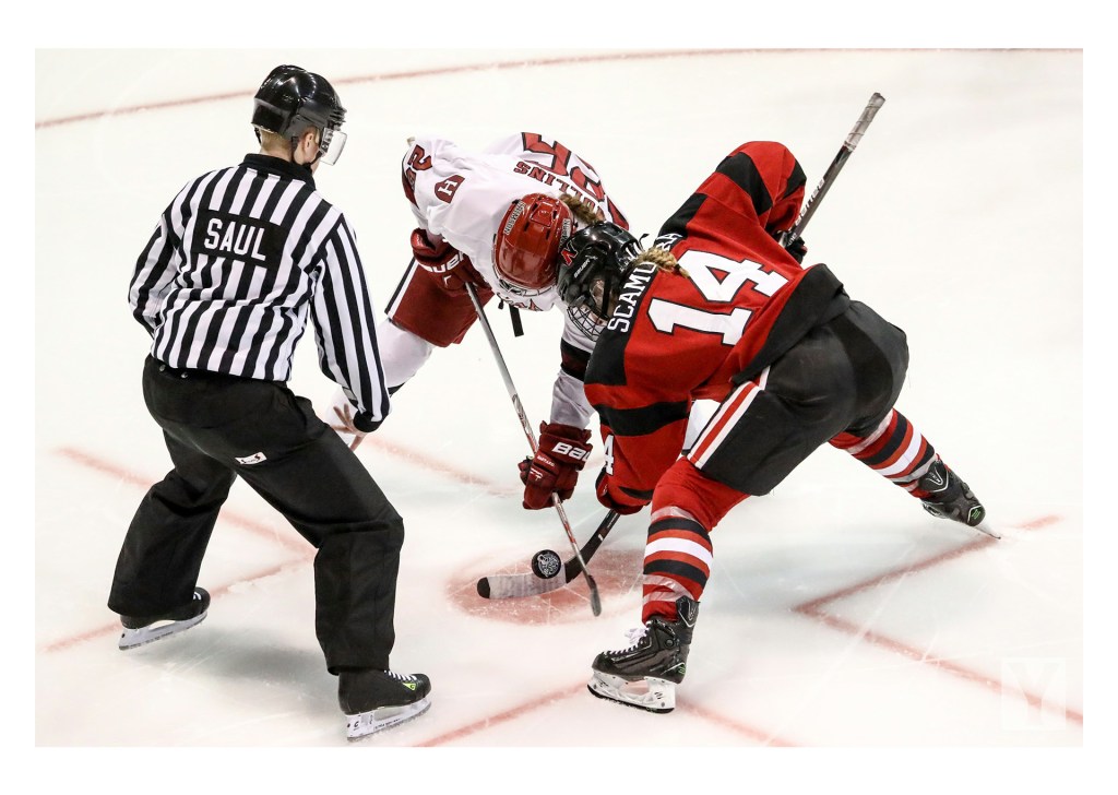 A close up picture of two female hockey players from opposite teams going for the hockey puck after it has just been dropped by the referee to signal the start of the game.