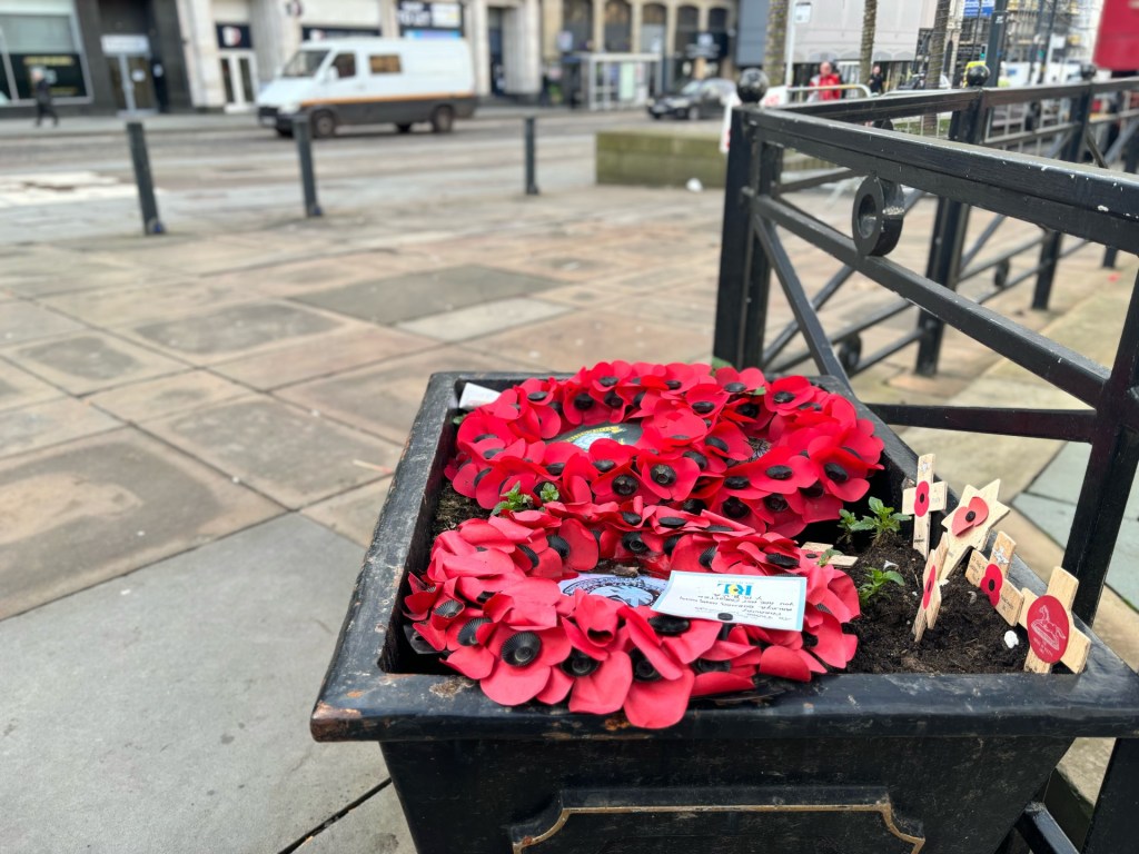 A poppy wreath in a pot of soil with crosses standing up in the soil and car passing by in the background that blurry but the main focus is on the wreaths
