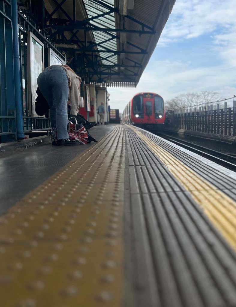 Taken on the tube station platform with the train arriving whilst a woman is going through her bag which is on the platform floor. 