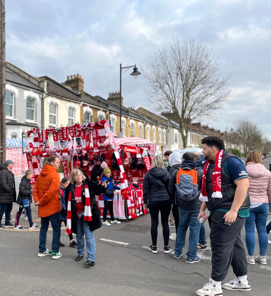 A woman stands in front of a Arsenal merchandise stall on situated directly opposite Arsenal tube station and on the left of the image as a group of people stand on the right. 
