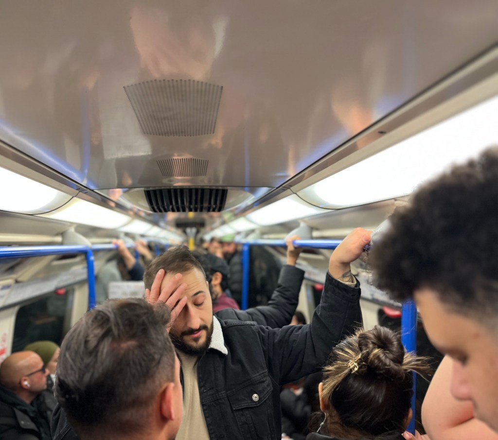 A man has his hands on his head as the rest of the tube carriage behind him is nearly at full capacity