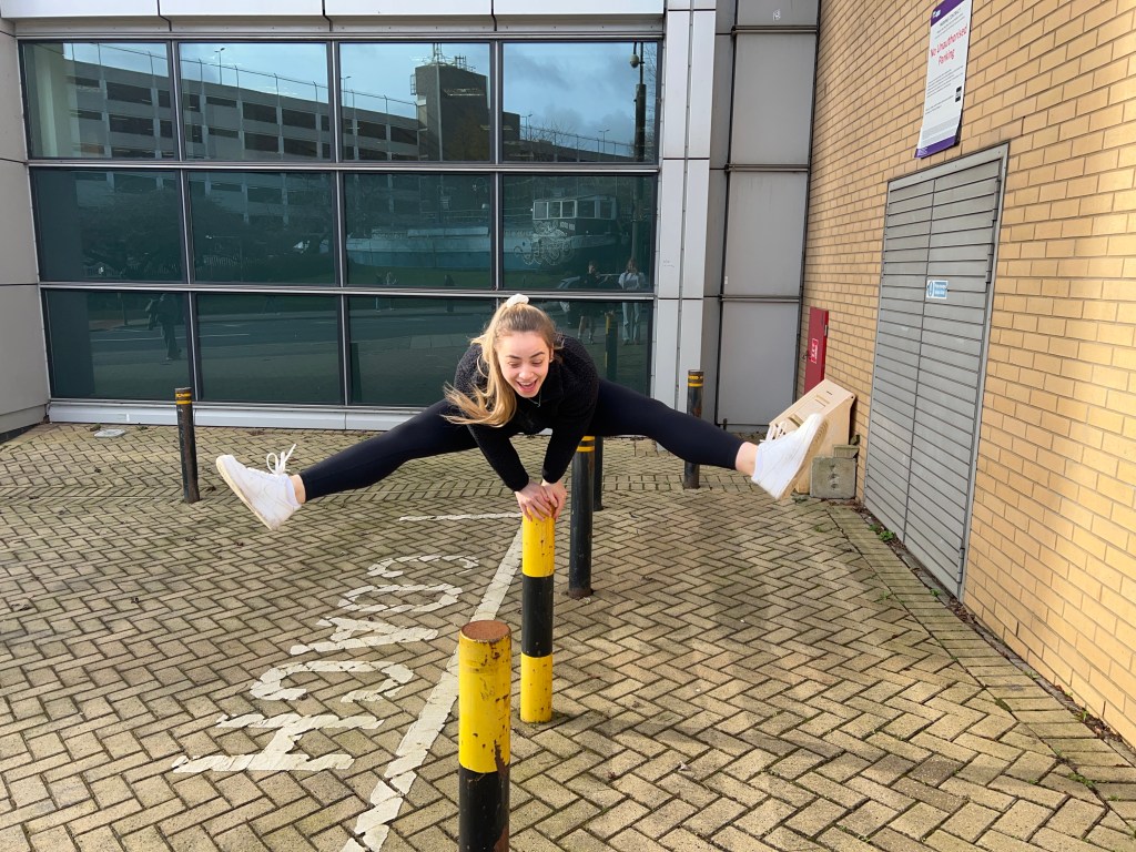 A girl is jumping with her legs out to the side over a black and yellow bollard