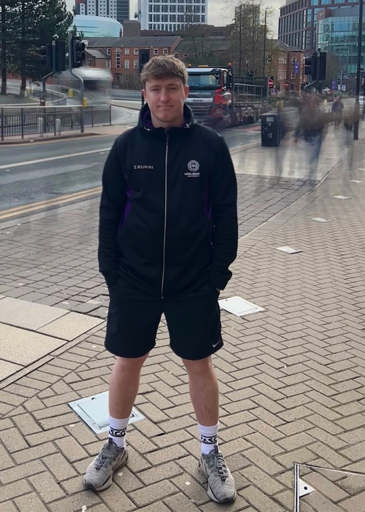 A photo of a male standing still in front of a busy background, with motion blurred behind him. The live photo long