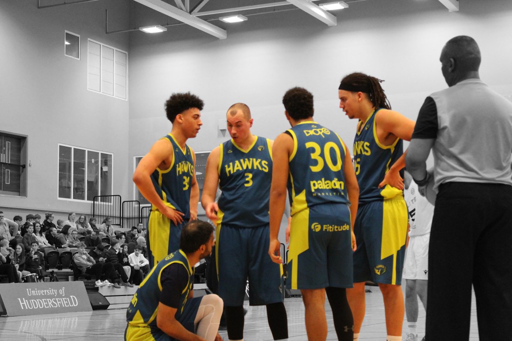 The West Yorkshire Hawks having a team talk on the court with a black and white background, whilst the players are in colour.