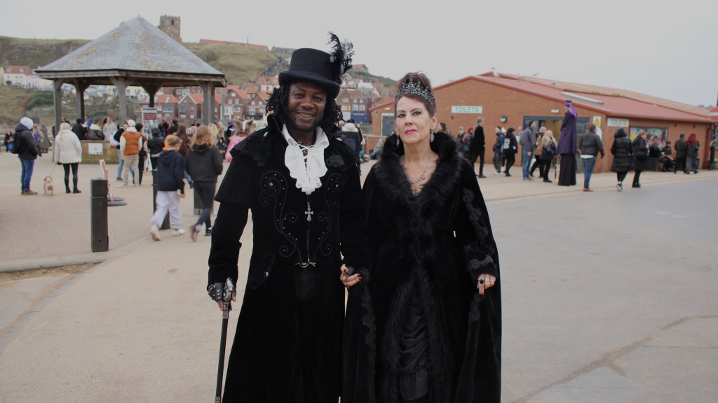 A couple linking arms in front of a bandstand. They are wearing Victorian outfits. The picture is over exposed.