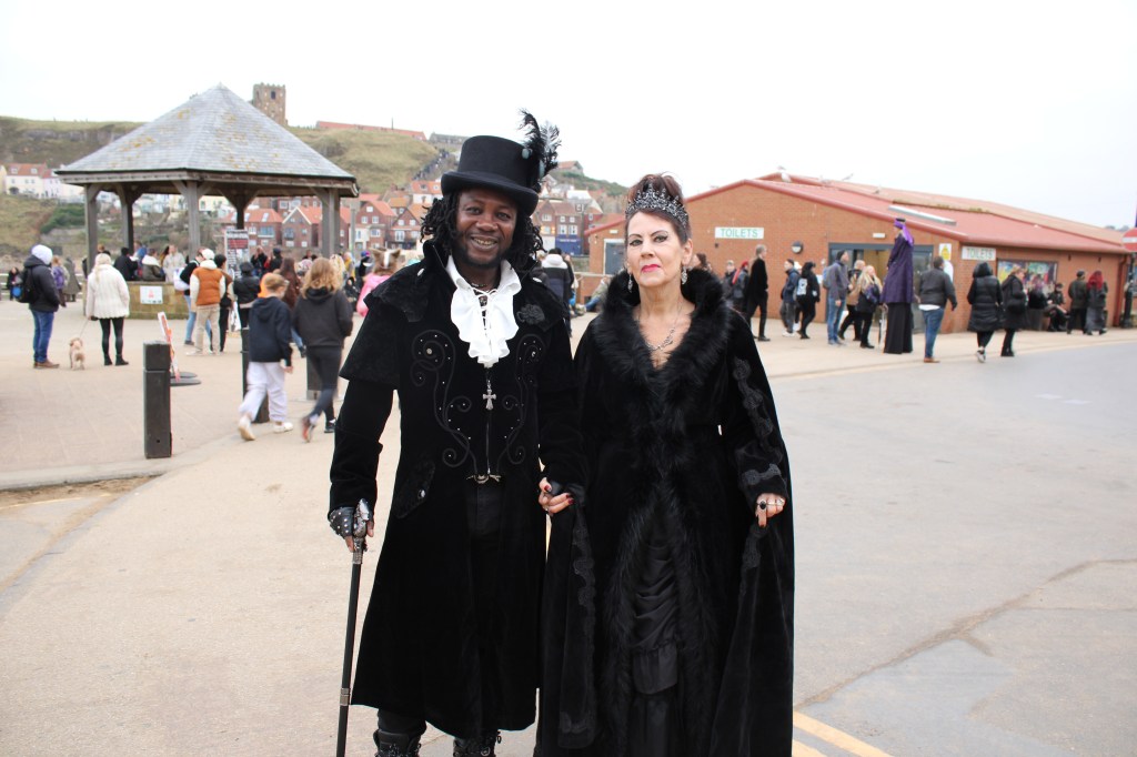A couple linking arms in front of a bandstand. They are wearing Victorian outfits. The picture is over exposed.
