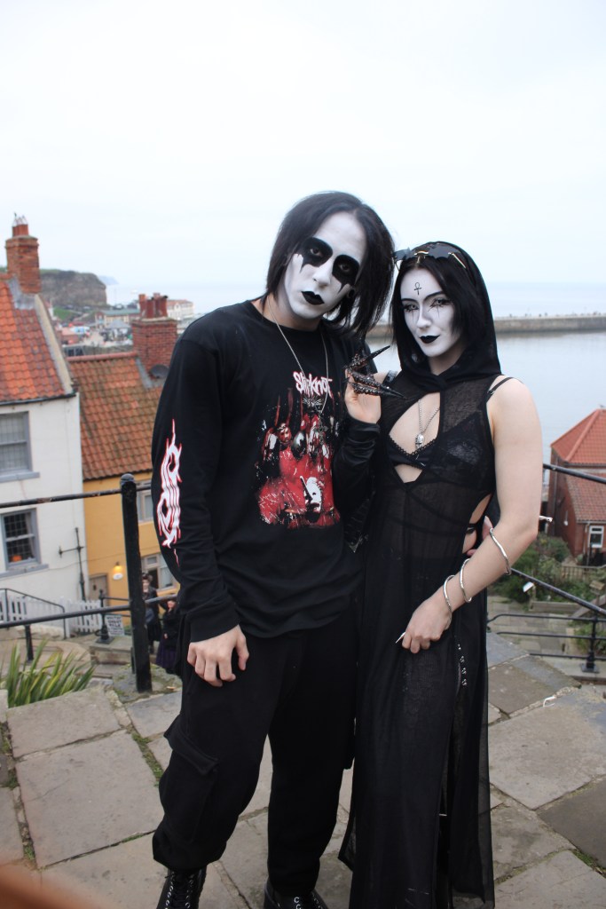 A couple arm in arm, both wear gothic outfits with black and white face paint with long black hair. The photo is in portrait.