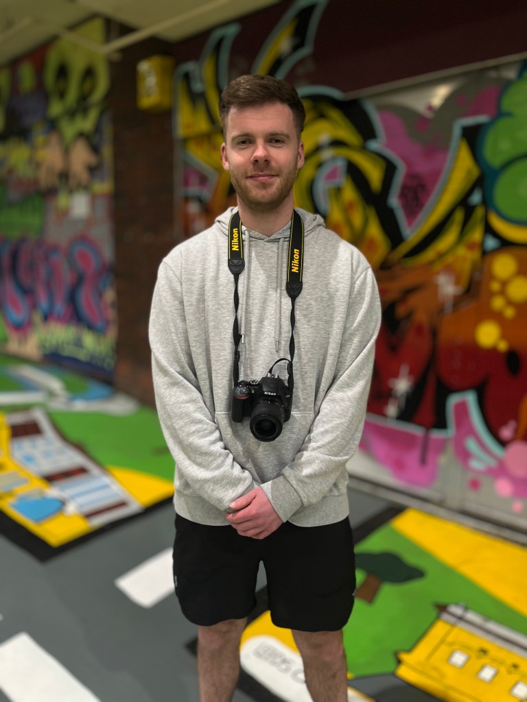 A young man with a camera around his neck grins while standing in front of a colorful graffiti mural inside Kirkgate Market