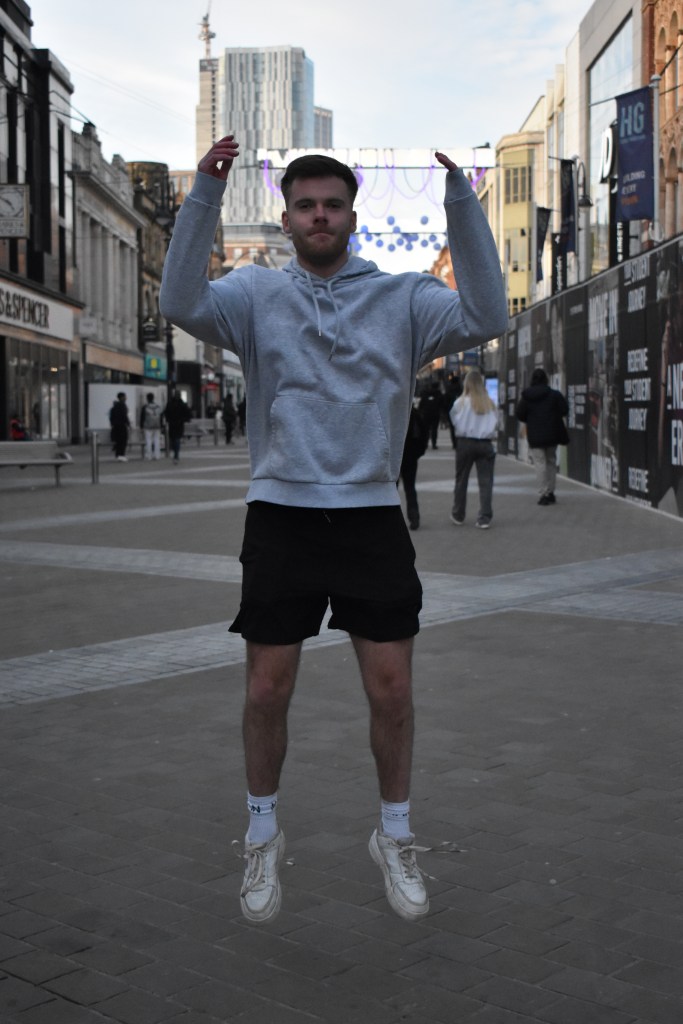 A man mid-jump, captured with a fast shutter speed, showing sharp details of his movement with minimal blur, frozen in mid-air. The background features a busy shopping strip