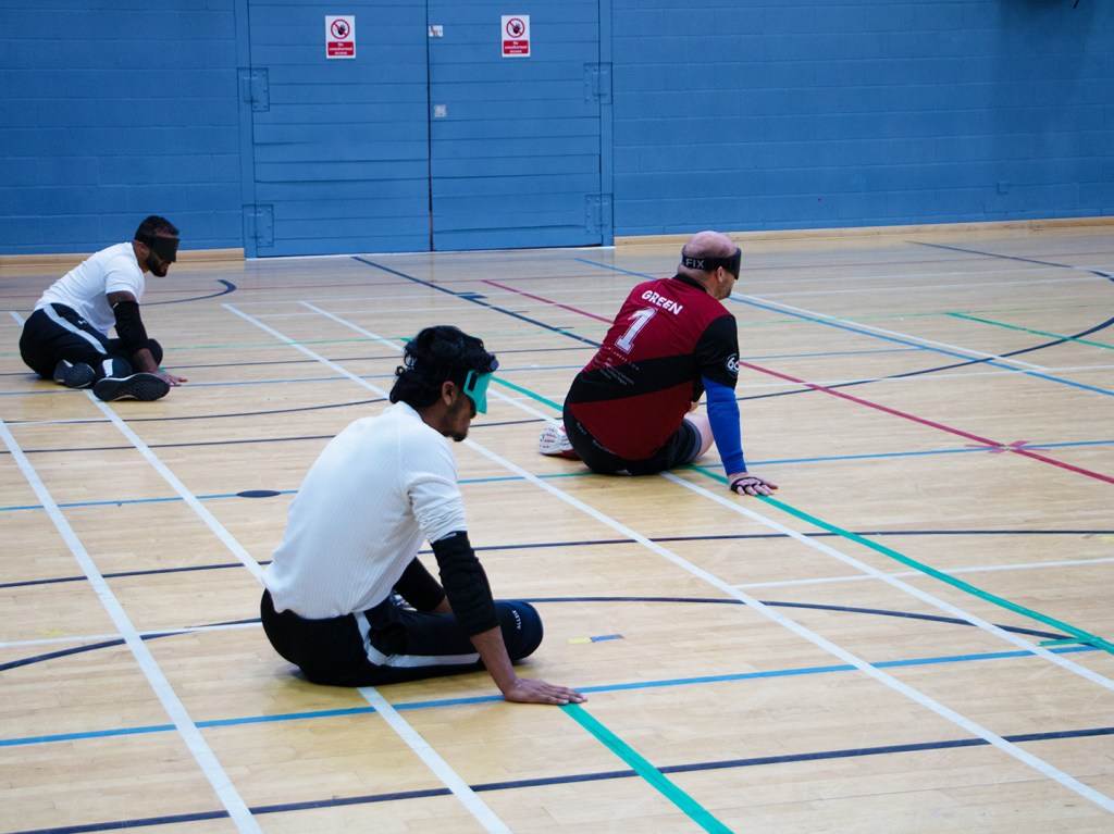 Three men sit in a triangualr formation on a gymnasium floor. They all have their hands on the floor and eye masks on