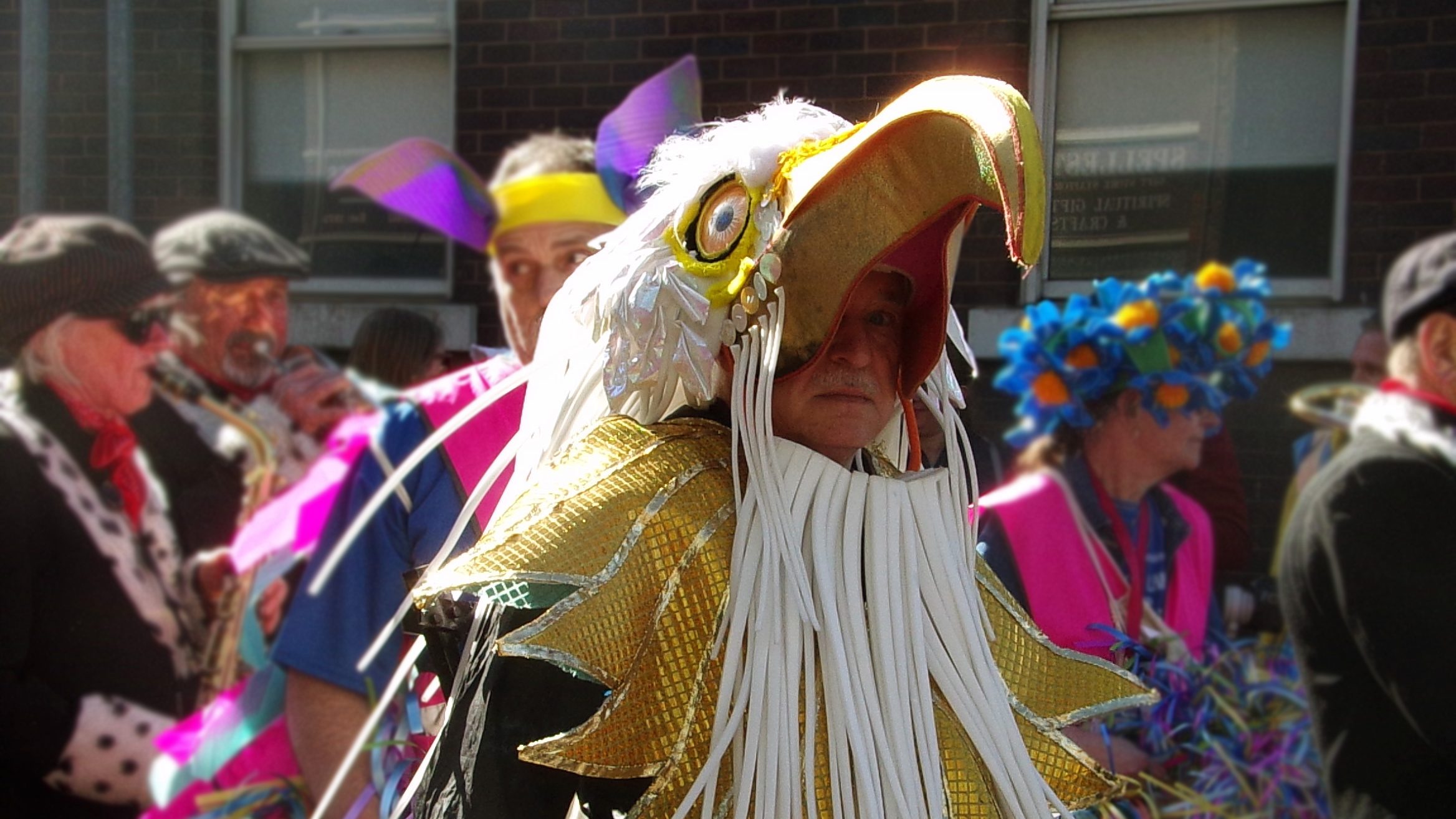A man wearing an eagle costume looks into the camera. The costume is a giant eagle's head which covers the top of his head. The golden beak shines into the camera.