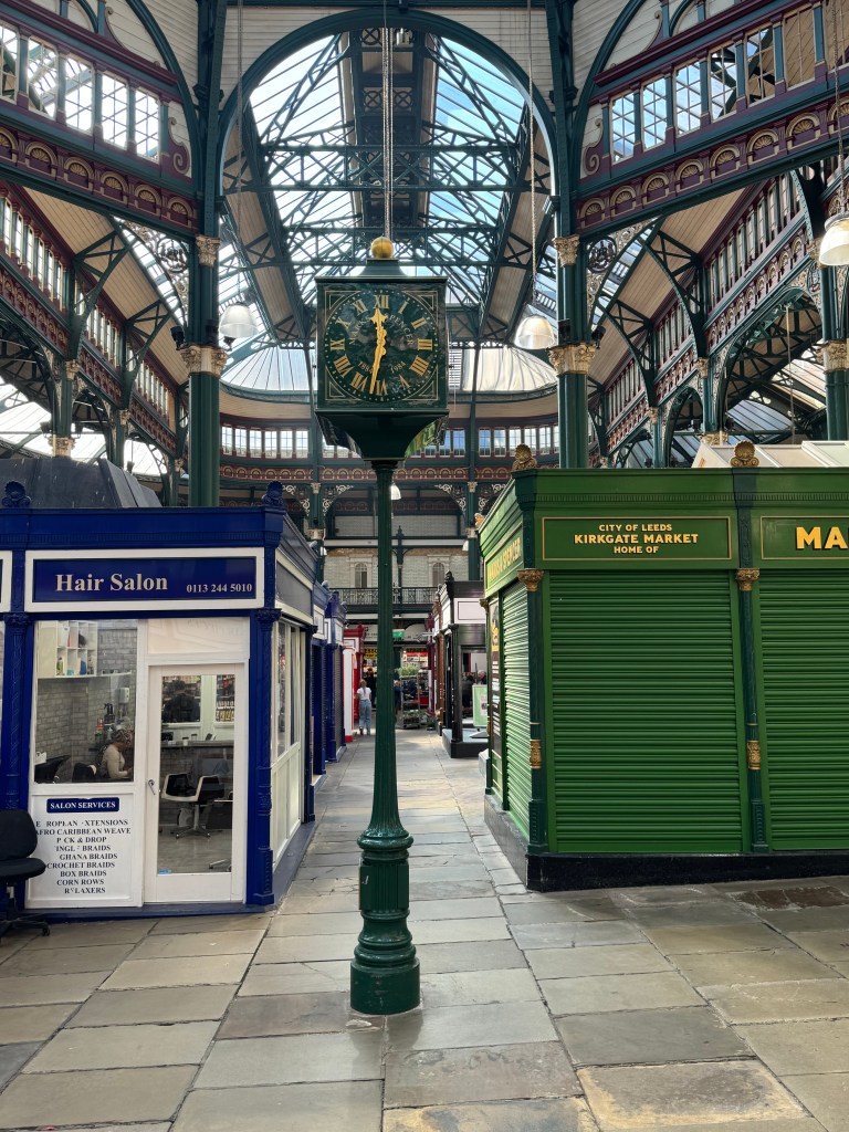 An old dark green clock with gold detailing stands proudly between the walkways of Kirkgate Market, with light streaming in from the roof above and stalls visible in the background