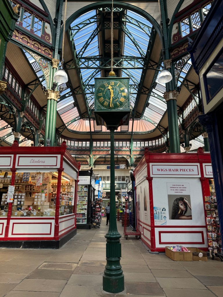 An old dark green clock with gold detailing stands proudly between the walkways of Kirkgate Market, with light streaming in from the roof above and stalls visible in the background