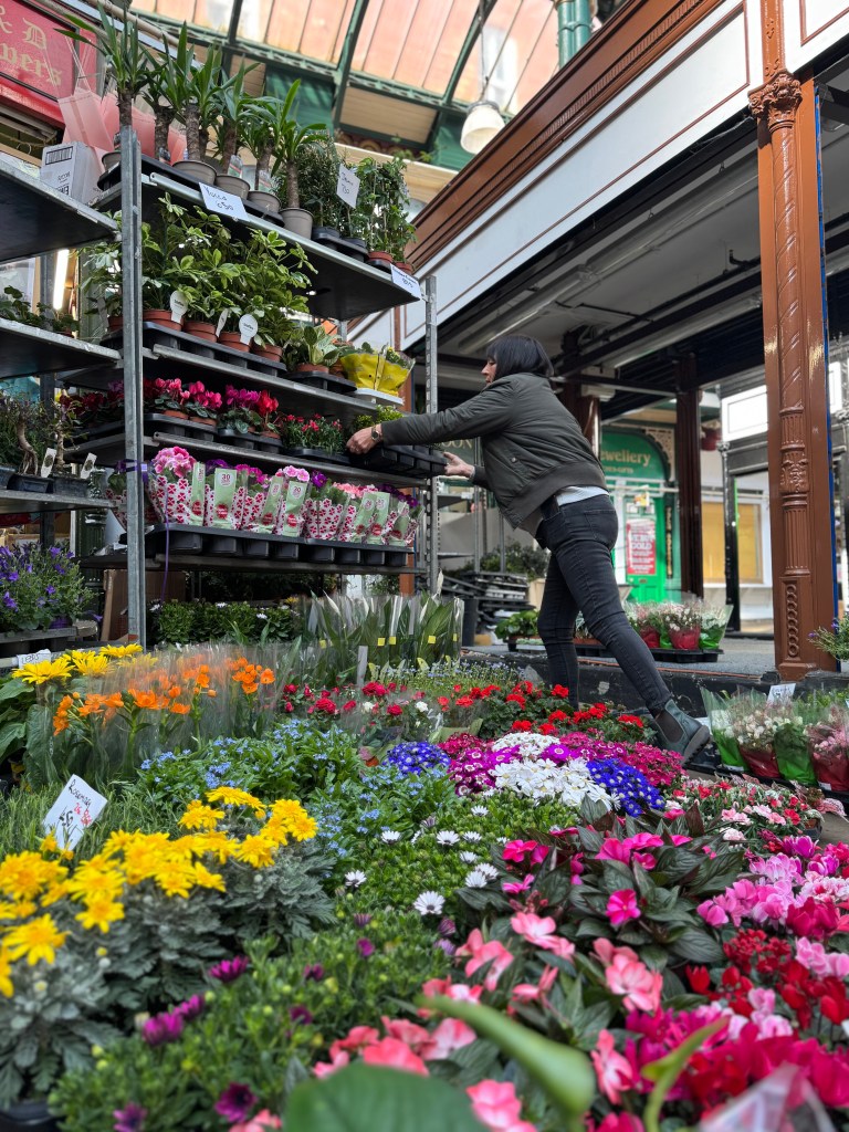A woman displays vibrant flowers at a stall inside Kirkgate Market, with natural light streaming in from the roof above