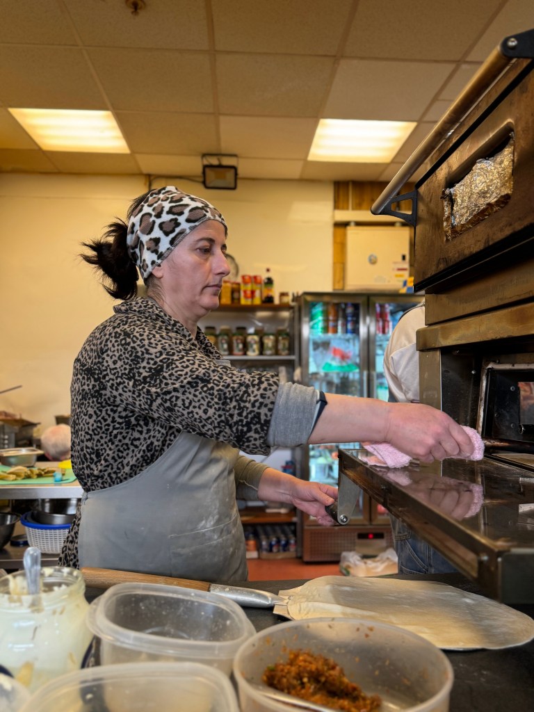 A woman wearing an apron and a cheetah print fleece with a matching bandana prepares flatbread at Eastern Oven, with ingredients in the foreground and the kitchen visible in the background