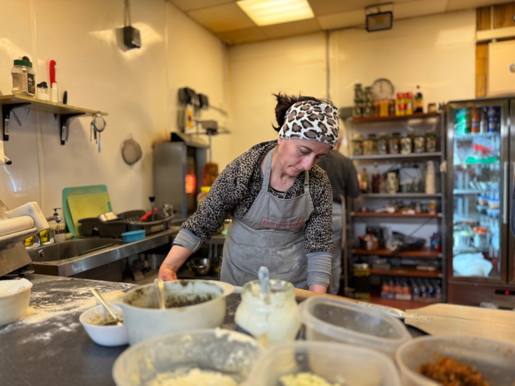 A woman wearing an apron and a cheetah print fleece with a matching bandana prepares flatbread at Eastern Oven, with ingredients in the foreground and the kitchen visible in the background