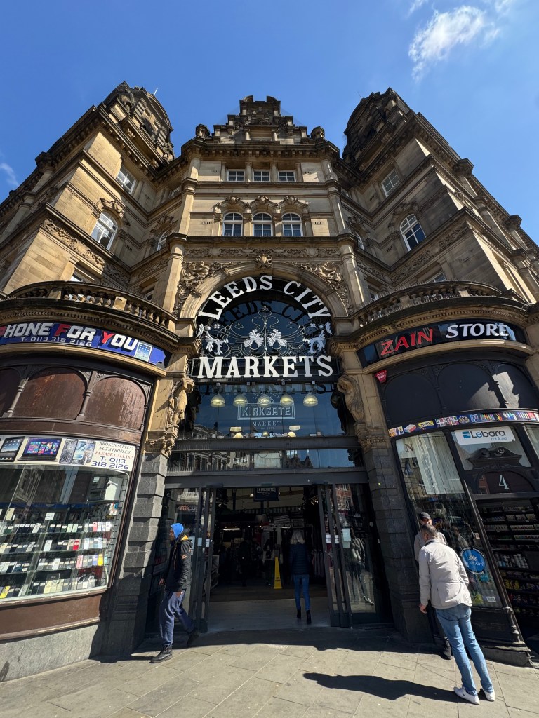 The main entrance of Kirkgate Market on a sunny day, with people walking in and out of the building
