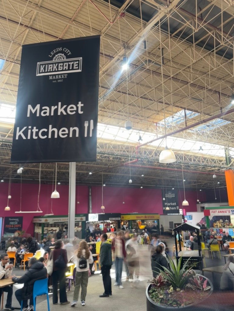 
The vibrant and busy Kirkgate Market Foodhall, with some people blurred to create a timelapse effect, and a "Market Kitchen" sign visible in the scene