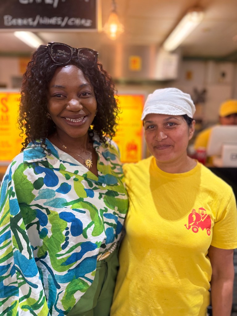 Jennifer Barry from Jenny’s Jerk Chicken and Manjit Kaur from Manjit’s Kitchen standing side by side in Kirkgate Market, smiling at the camera with their arms around each other’s backs