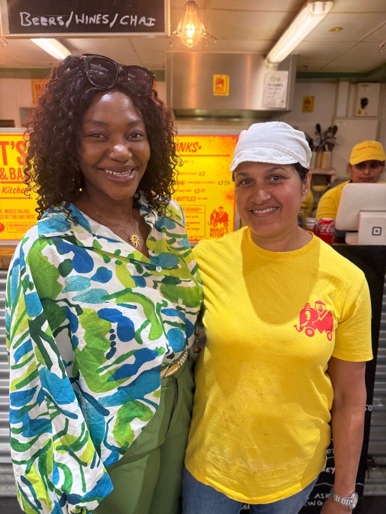 Jennifer Barry from Jenny’s Jerk Chicken and Manjit Kaur from Manjit’s Kitchen standing side by side in Kirkgate Market, smiling at the camera with their arms around each other’s backs