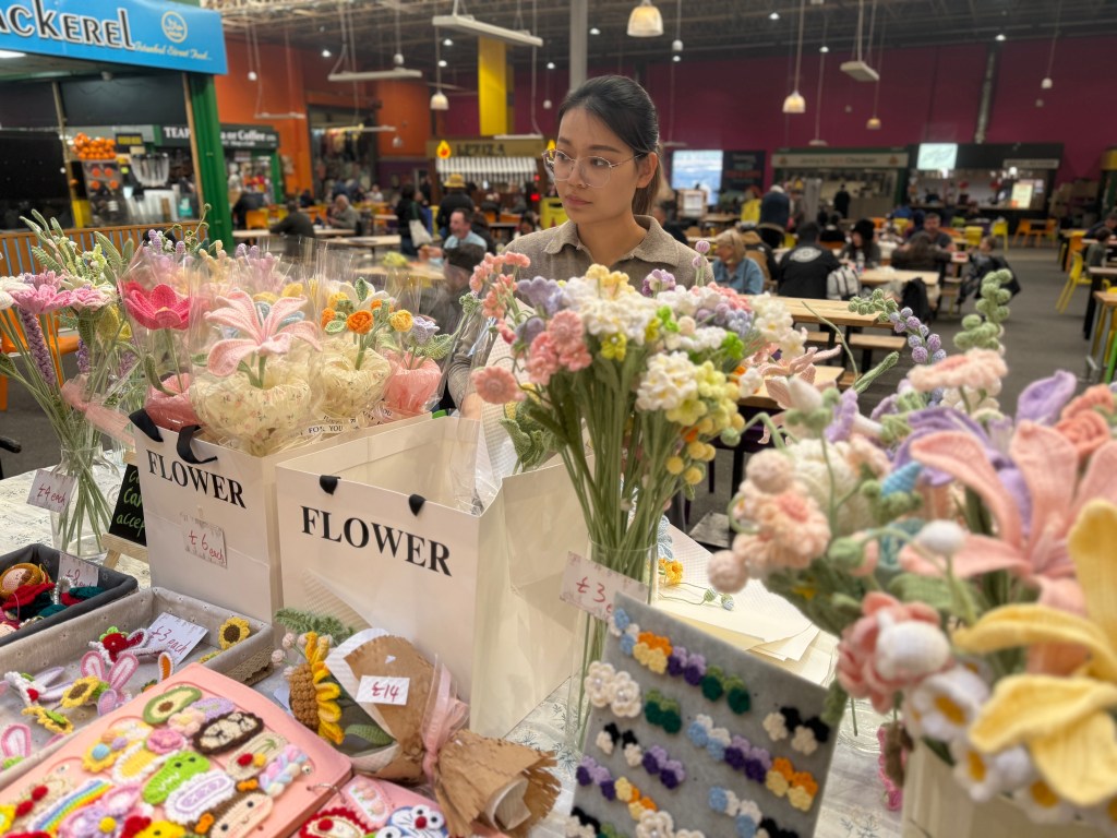 A woman stands behind her cart in the centre of Kirkgate Market’s Foodhall, speaking with a customer. The cart is filled with colourful knitted flowers and gifts
