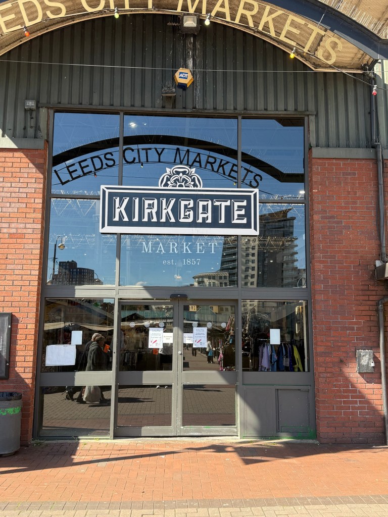 The back entrance of Kirkgate Market with glass automatic sliding doors and a black sign with white lettering reading "Kirkgate Markets." Reflected in the glass is partial text from a sign above that reads "Leeds City Markets." The scene is lit by bright natural light