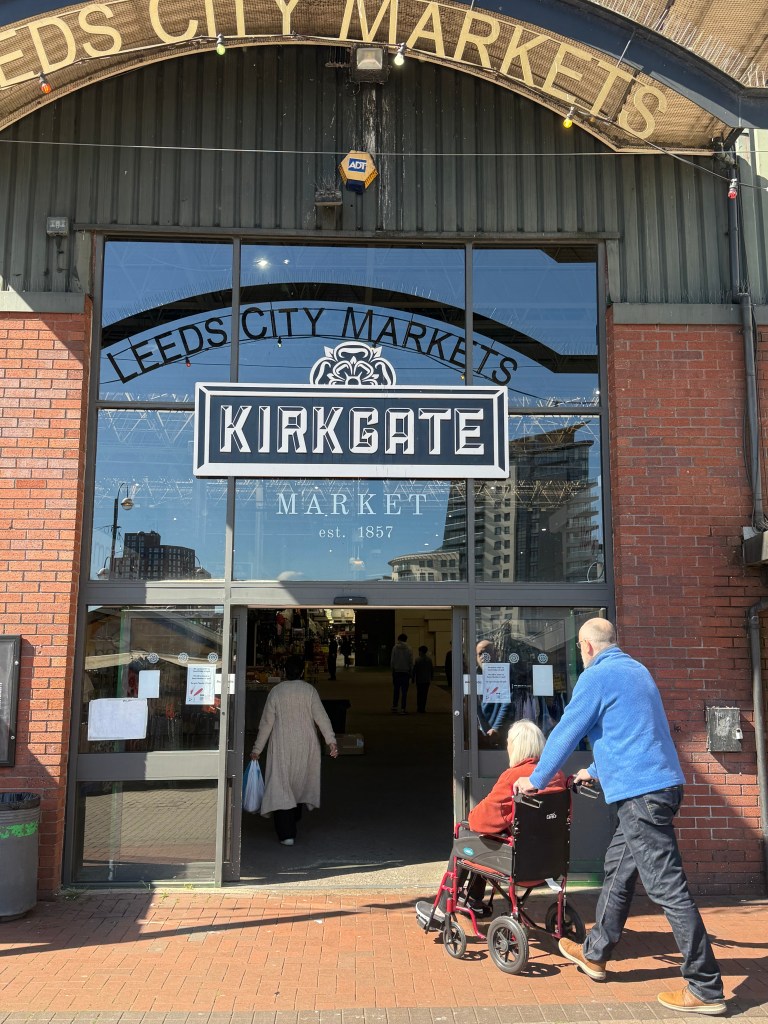 A man wheels a person in a wheelchair through the back entrance of Kirkgate Market, which features glass automatic sliding doors and a black sign with white lettering reading "Kirkgate Markets." Reflected in the glass is partial text from a sign above that reads "Leeds City Markets." The scene is lit by bright natural light
