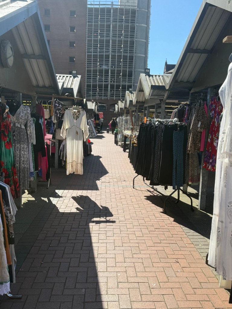 The walkways outside Kirkgate Market, lined with stalls displaying clothes. The scene is bright with sunlight and a clear sky