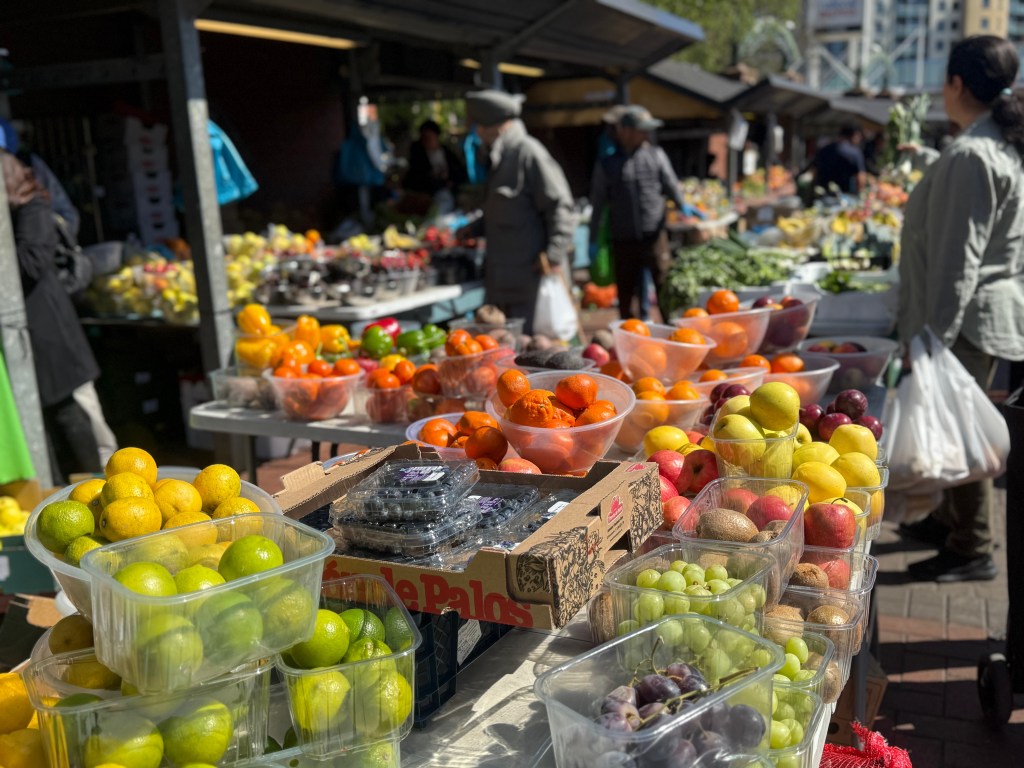 A colourful display of fruits outside Kirkgate Market, including grapes, limes, kiwis, blueberries, and oranges, with a stallholder tending to a customer in the blurred background
