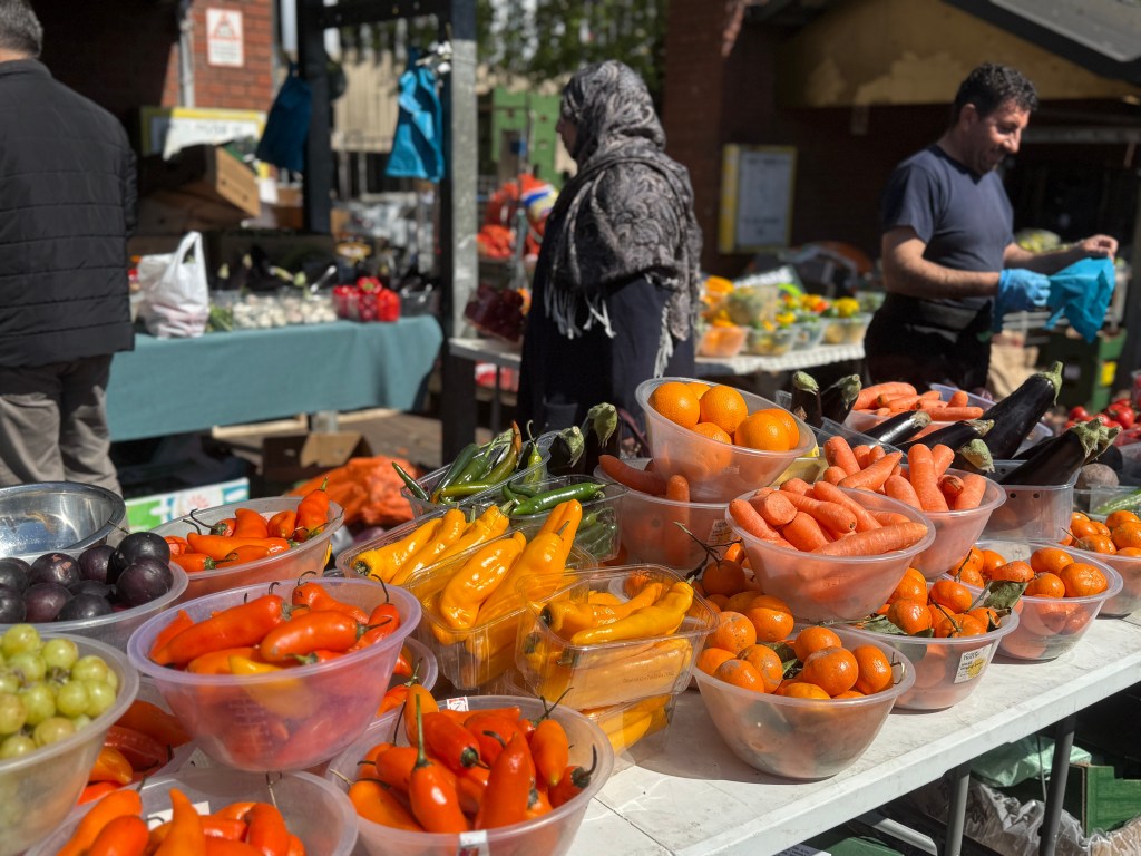 A colourful display of peppers, aubergines, carrots, grapes, and oranges outside Kirkgate Market on a sunny day. A woman in a hijab stands in the background, while the male stallholder appears to be smiling. The background is slightly out of focus