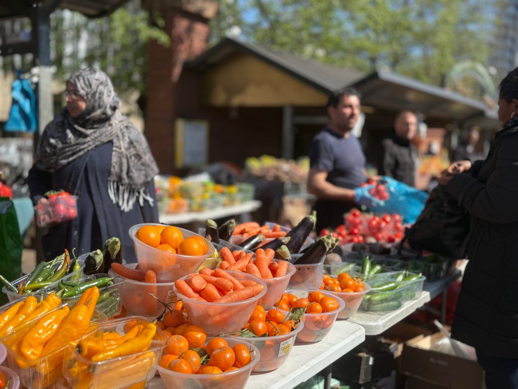 A colourful display of peppers, aubergines, carrots, grapes, and oranges outside Kirkgate Market. The produce in the foreground is in sharp focus, while a woman in a hijab holding a punnet of strawberries and a male stallholder to her right are slightly blurred in the background