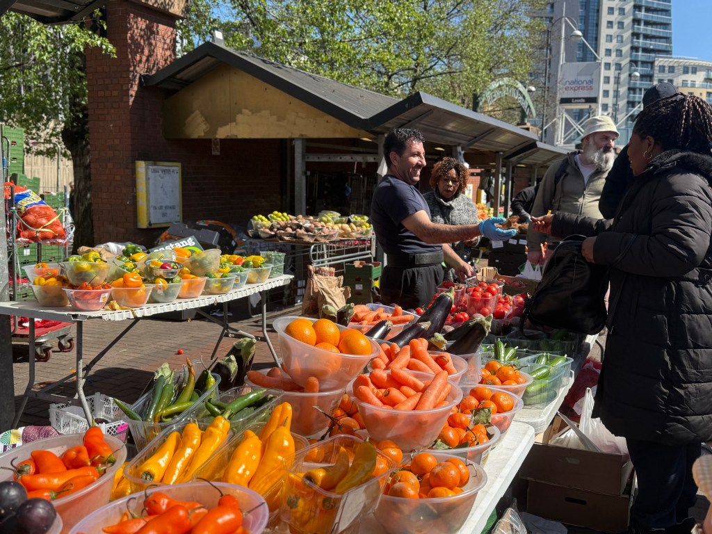 A vibrant display of peppers, carrots, grapes, and oranges outside Kirkgate Market. The produce is in sharp focus in the foreground, while a vendor with his hand outstretched laughs with a group of customers on the right side of the shot