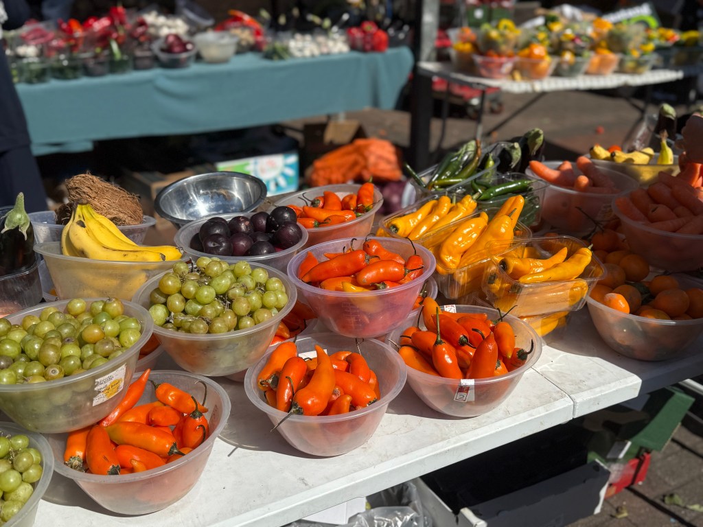 A vibrant display of peppers, carrots, grapes, and oranges at Kirkgate Market. The produce is in sharp focus, showcasing its bright colours