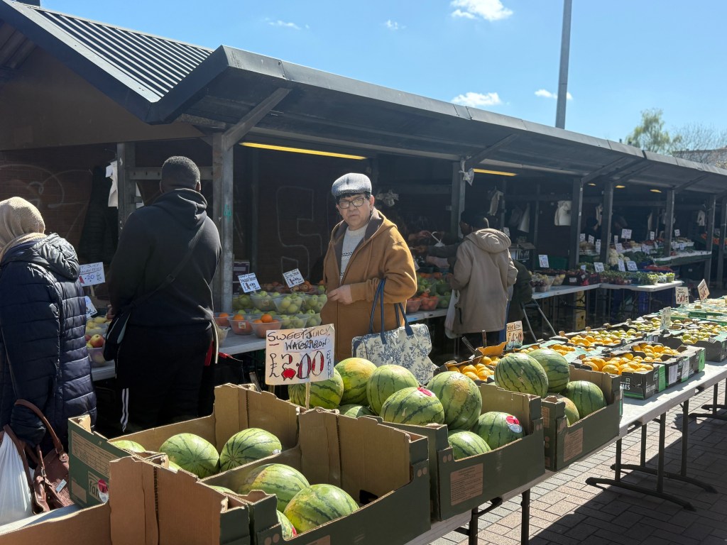 Watermelons in crates on tables in the foreground, with sheltered stalls behind and customers walking along the walkway, looking at the market offerings. The sky is clear and bright blue