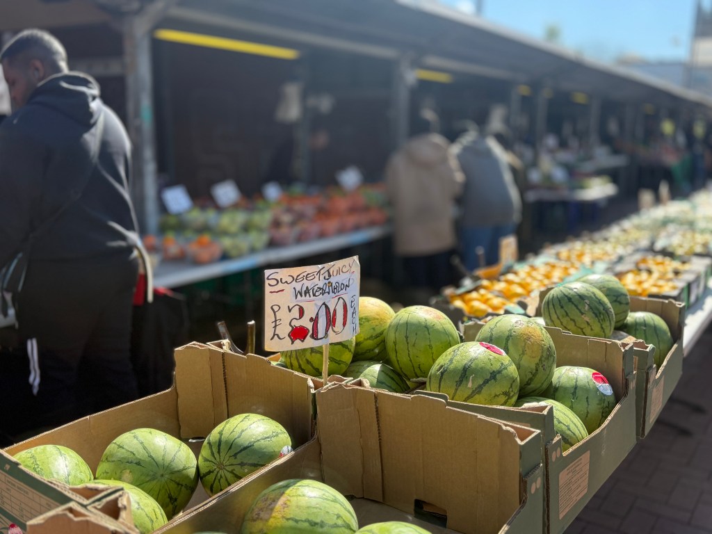 Watermelons in crates on tables in sharp focus, with a blurred background of sheltered stalls and customers walking along the walkway. The sky is clear and bright blue