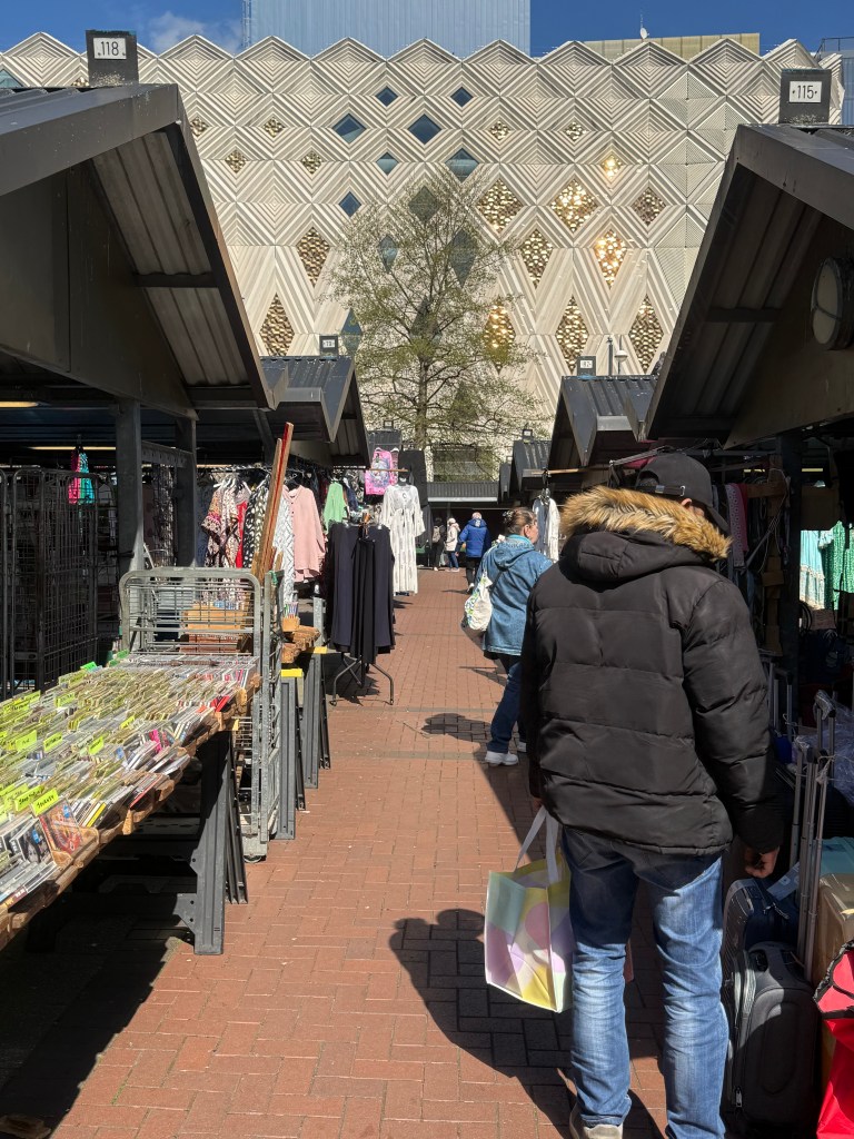 The walkways outside Kirkgate Market, lined with stalls displaying clothes. The old John Lewis building is visible in the background. The scene is bright with sunlight and a clear, blue sky
