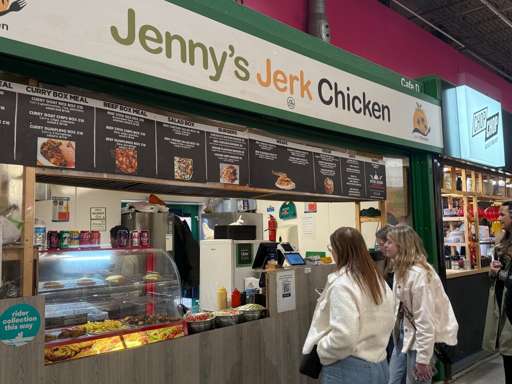 Jenny's Jerk Chicken food stall inside Kirkgate Market with a green painted front. Images of the menu items, including curry box meals, beef box meals, salad boxes, burgers, and wraps, are displayed around the opening hatch. Customers are visible near the stall, and someone can be seen working in the kitchen behind the counter