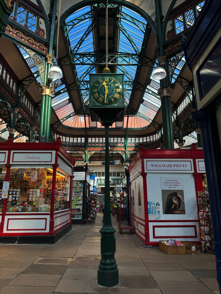 An old dark green clock with gold detailing stands proudly between the walkways of Kirkgate Market, with light streaming in from the roof above and stalls visible in the background