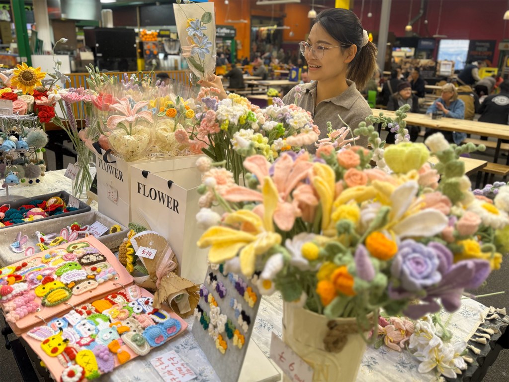 A woman stands behind her cart in the centre of Kirkgate Market’s Foodhall, speaking with a customer. The cart is filled with colourful knitted flowers and gifts