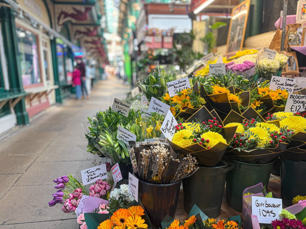 A close-up of a vibrant flower display in Kirkgate Market, with various colourful flowers in sharp focus and a blurred walkway in the background
