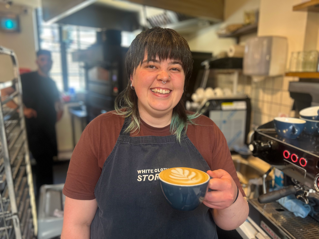 A barista holds a latte in a cafe, with the coffee machine next to her and a chef in the background